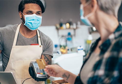 a customer interacts with a retail worker, both wearing masks, at a checkout station while scanning a credit card to pay